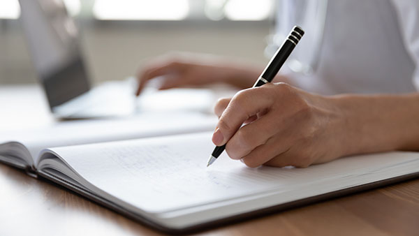 Close up of a person's hand holding a pen, and writing on a piece of paper laid on a desk
